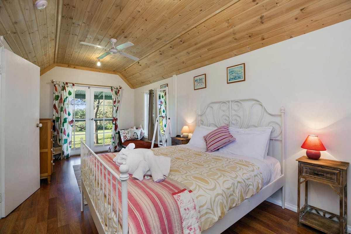 Un dormitorio con techo de madera, cama con ropa de cama estampada, mesita de noche con lámpara y vistas al exterior a través de una puerta.