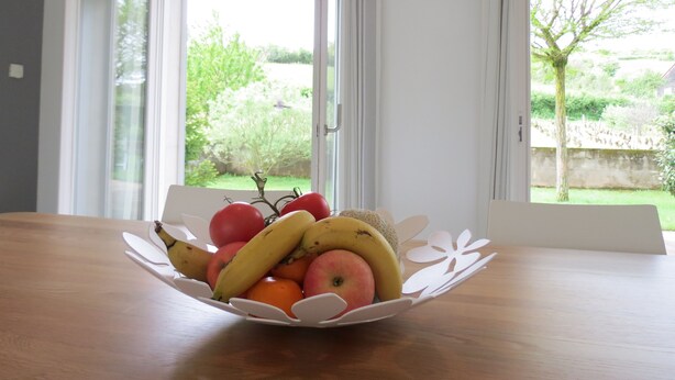 A fruit bowl on a table with a view of a garden through a window.