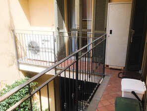 Een balkon met een metalen hekwerk, een witte prullenbak en een groene tas op de vloer.