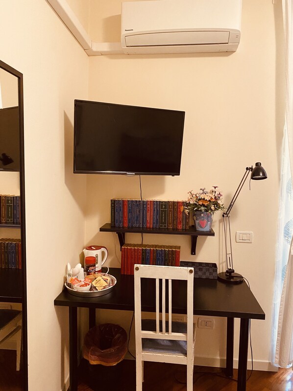 Una habitación con una televisión de pantalla plana montada en la pared, un escritorio con una silla, una estantería y una mesita con una taza de café y libros.