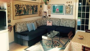 A living room with a sofa, a glass coffee table, and various decorative items including framed pictures and pottery on shelves.