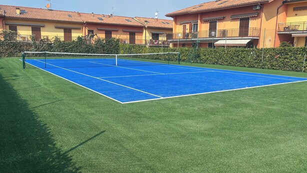 Una cancha de tenis al aire libre con red, rodeada de setos y edificios residenciales al fondo.