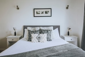Een net opgemaakt bed met een witte deken en een grijze gebreide plaid. Twee nachtkastjes met kleine vazen erop.
