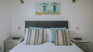 A bed with a striped bedspread, two bedside tables, and a wall-mounted painting of a house and bicycle. - Atlantic Reach Cottage, Newquay.  Large 4 bed villa with leisure services & WiFi (Newquay)