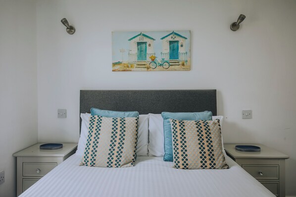A bed with a striped bedspread, two bedside tables, and a wall-mounted painting of a house and bicycle. - Atlantic Reach Cottage, Newquay. Large 4 bed villa with leisure services & WiFi (Newquay)