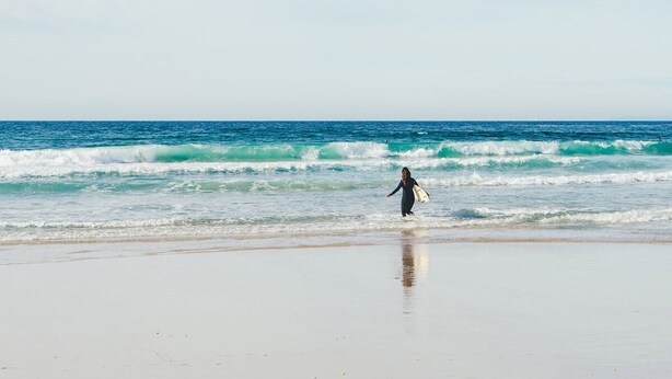A beach with a person walking along the shore carrying a surfboard. - Atlantic Reach Cottage, Newquay.  Large 4 bed villa with leisure services & WiFi (Newquay)