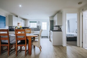 Een moderne keuken met witte kastjes, een eettafel met stoelen en uitzicht op een slaapkamer met een bed en gordijnen.
