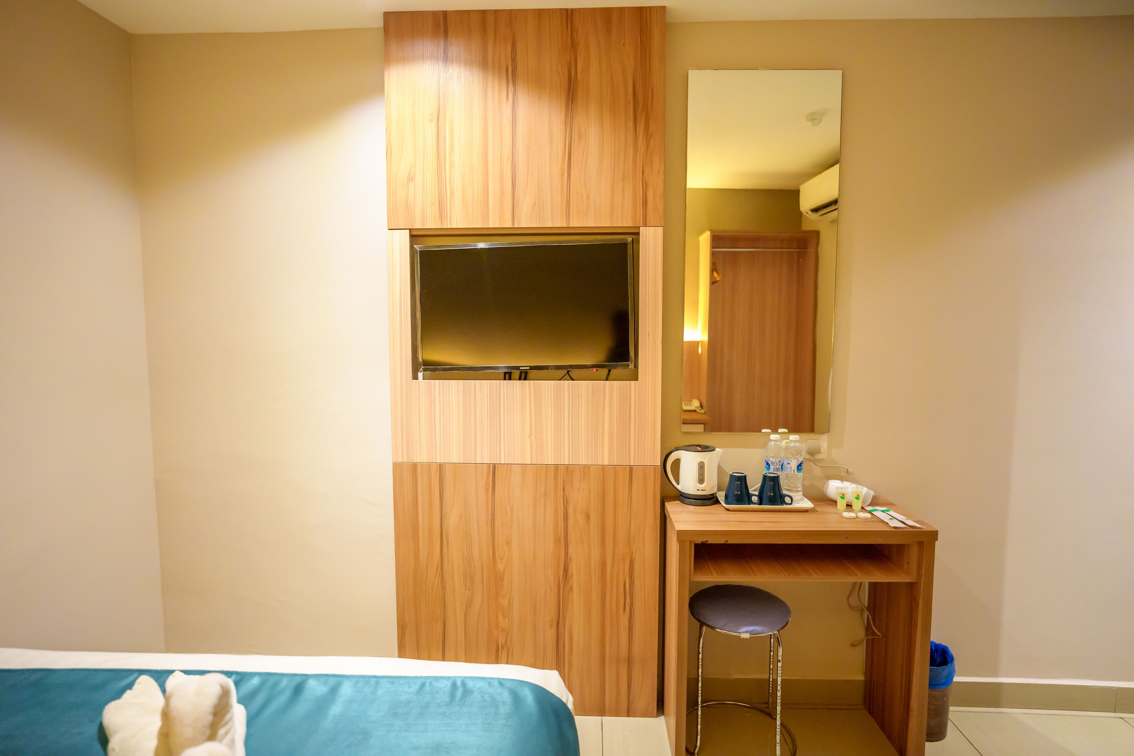 A hotel room with a bed, a wooden desk, a mirror, a television, and a small stool.