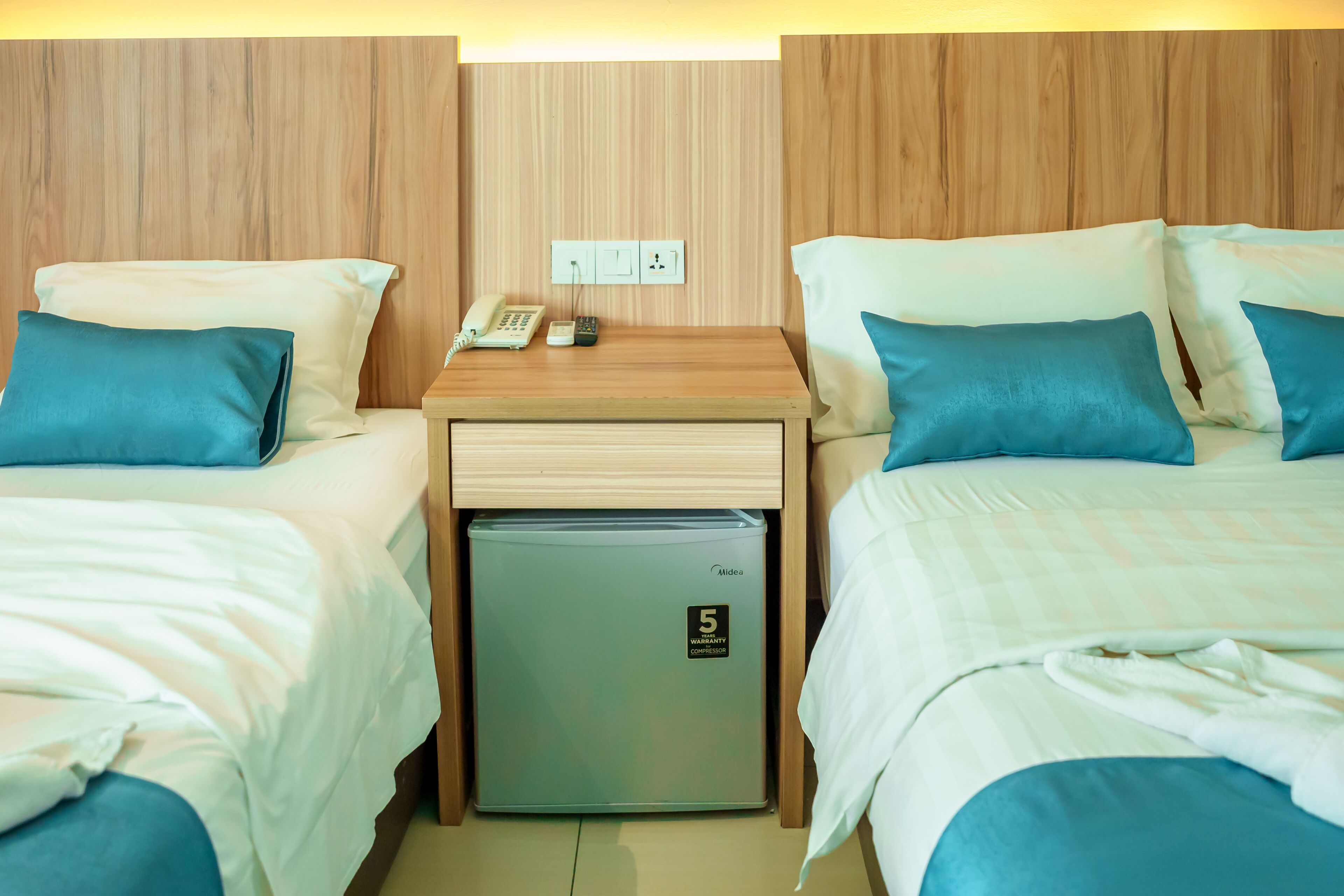 Habitación de hotel con dos camas, cada una con almohada azul, una mesita de noche de madera con teléfono y un mini refrigerador, y un cabecero de madera.