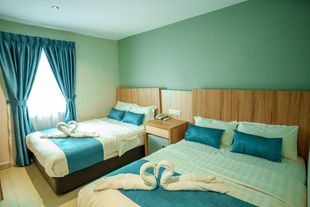 Habitación de hotel con dos camas, cada una con cubrecama azul y almohadas blancas. Hay dos mesitas de noche con lámparas, un cabecero de madera y una ventana con cortinas azules.