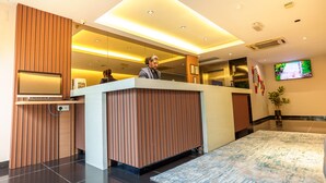 A modern reception area with a wooden reception desk, a flat-screen TV mounted on the wall, and a potted plant. - Alaxis Hotel (Kuala Lumpur)