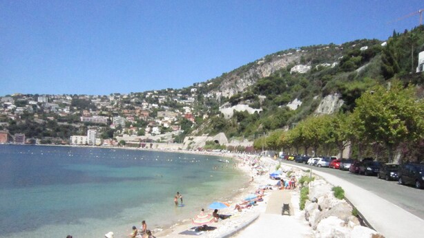 A coastal beach with people sunbathing and swimming, a clear blue sea, and buildings in the background. - Residence of the Guardian Angel, Beachfront apartment, Villefranche / Mer (Villefranche-sur-Mer)