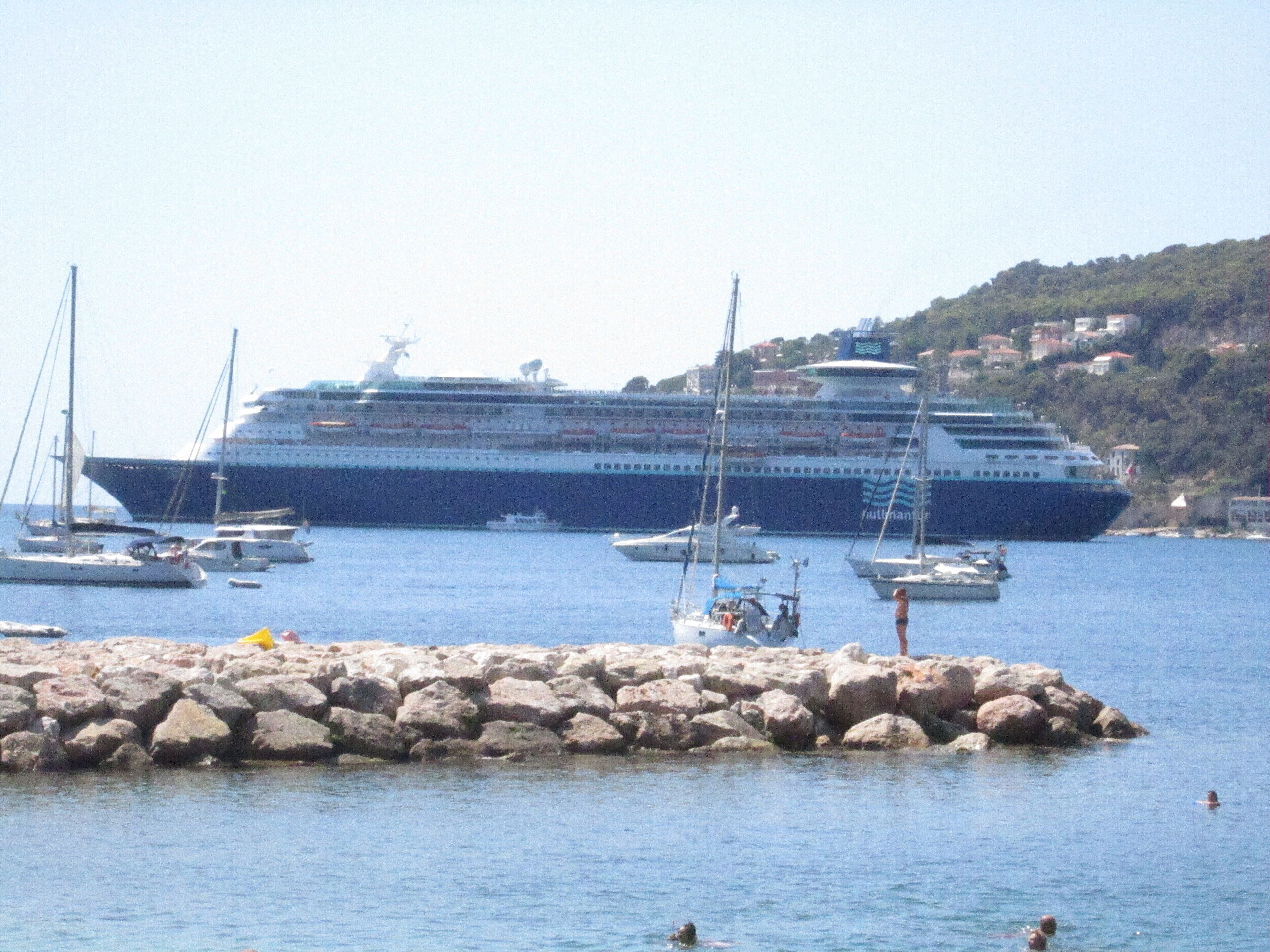 A cruise ship docked at a marina with several smaller boats and yachts around it.
