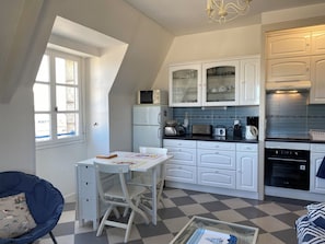 Cocina funcional con armarios blancos, microondas y estufa. Hay una pequeña mesa de comedor con sillas y una silla azul con cojín.