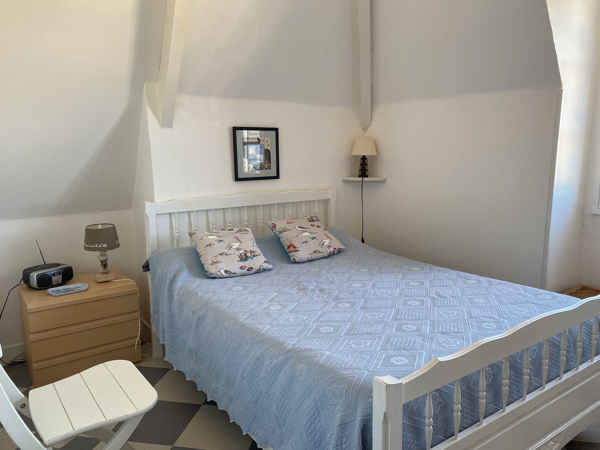 Un dormitorio con una cama, una mesita de noche con lámpara, una silla y una pequeña mesa con un radio.