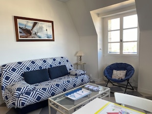 Una sala de estar con un sofá estampado, una mesa de centro de vidrio, una silla, una lámpara y un ventanal con vistas.
