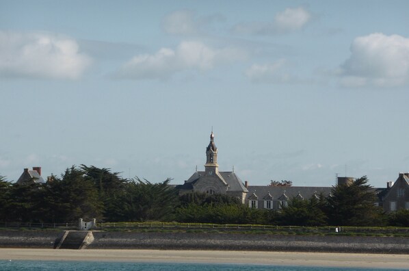 Un edificio costero con una torre destacada, rodeado de árboles y una playa en primer plano.