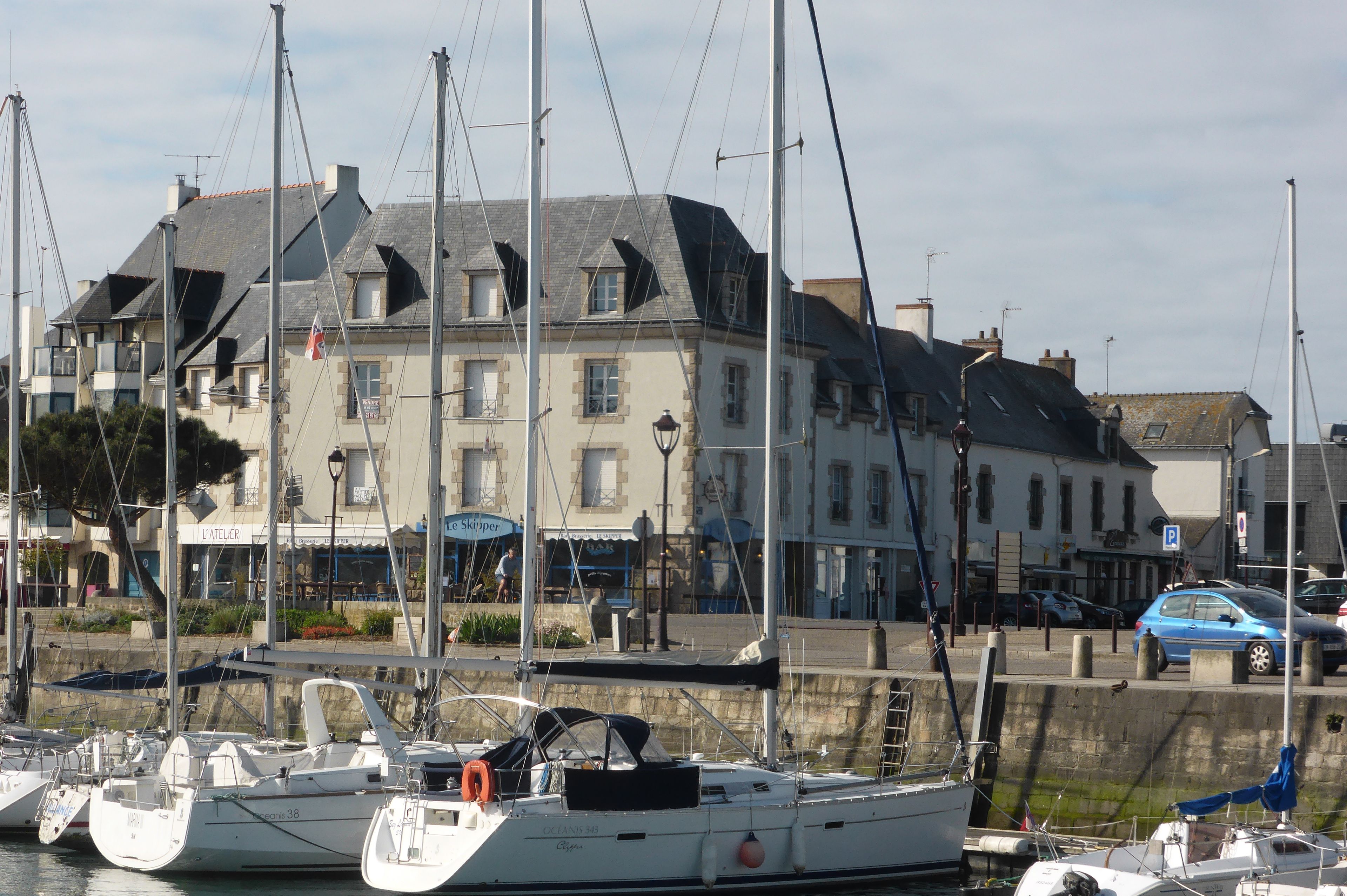 A marina with several sailboats docked. There is a building with a sign that reads "Le Skipper" and another sign that says "L'Atelier".