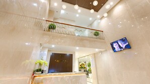 Un moderno vestíbulo de hotel con un mostrador de recepción de mármol, un mostrador de recepción de madera, un balcón con plantas y un televisor montado en la pared.