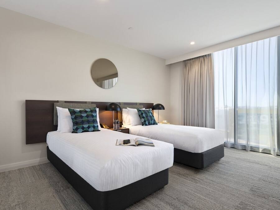 A hotel room with two beds, a large window with curtains, a round mirror on the wall, and a small table with a lamp.