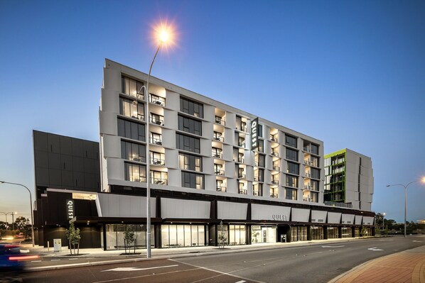 Un hôtel moderne à plusieurs étages, doté de grandes fenêtres et d’une enseigne « Quest » bien visible.