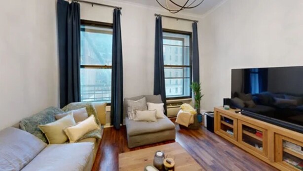 A living room with a sofa, armchair, and ottoman, a television on a wooden stand, and large windows with curtains. - Central Park, Theater District & Columbus Circle. New Renovation 3 Bed / 2 Bath. (New York City)