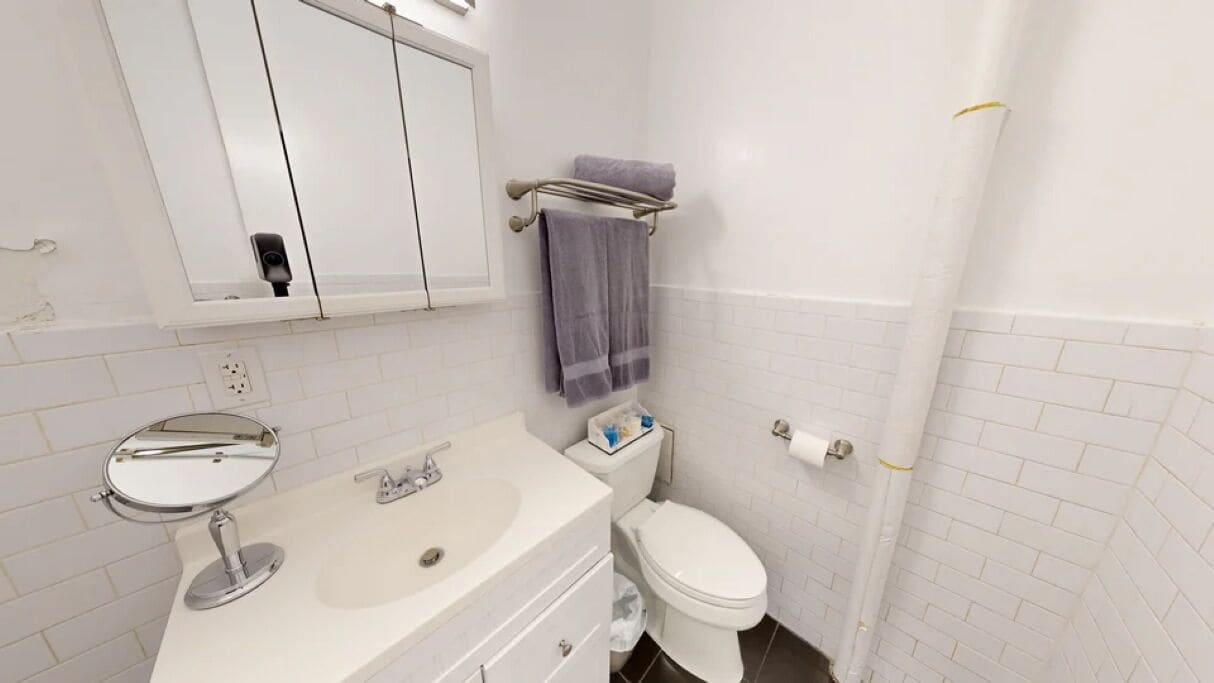 A bathroom with a white sink, mirror, and toilet. There is a towel rack with a towel and a roll of toilet paper on the wall.