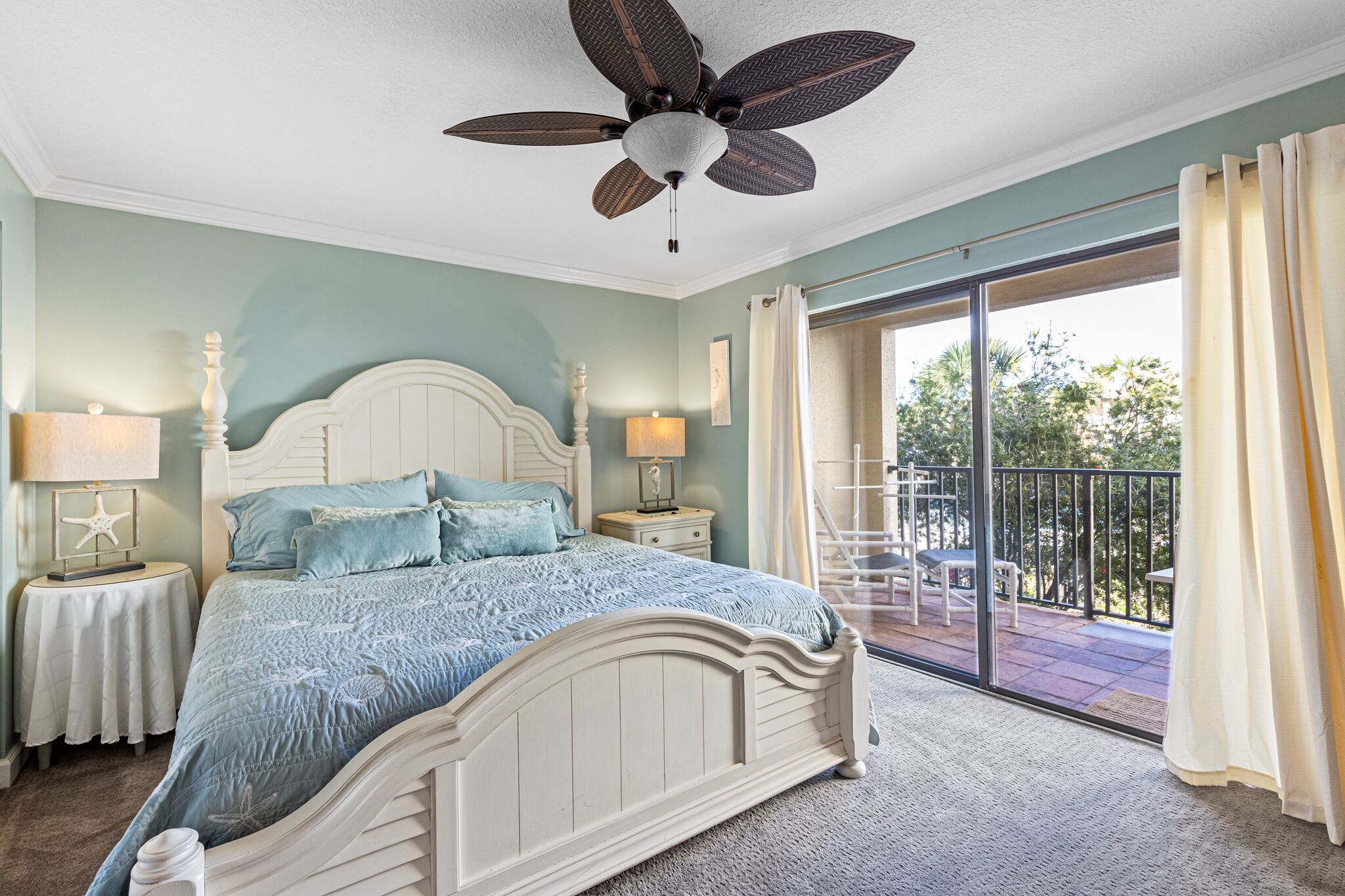 Une chambre à coucher avec un lit, un ventilateur de plafond, une table de chevet avec une lampe et une vue sur le balcon extérieur.