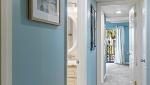 A hallway with light blue walls, a framed picture, and a door leading to a balcony with a view of palm trees. -  3 Bedroom Condo/ Sleeps 8 / Beautifully Upgraded (St Augustine)