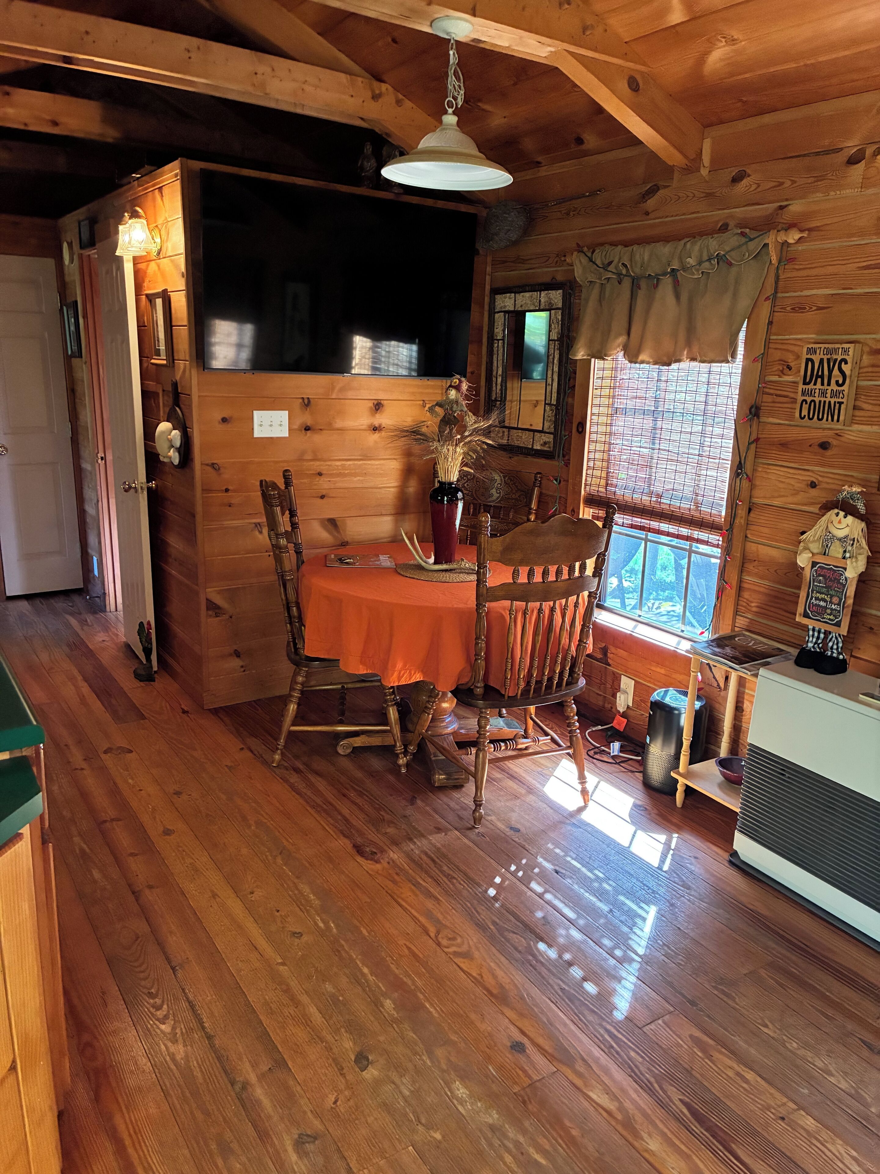Interior acogedor de una cabaña de madera con una mesa de comedor, sillas, un florero con plantas secas, una lámpara colgante y una ventana con vistas.