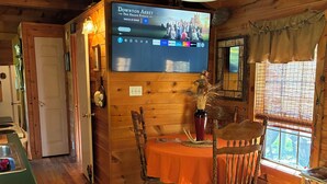 Ein Holzblockhaus-Interieur mit Essbereich, einem Fernseher, der ein Programm zeigt, und einem Fenster mit Vorhängen.