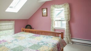Ein Schlafzimmer mit schrägen Wänden, einem hölzernen Bett, einem Fenster mit floralen Vorhängen und einem Oberlicht.