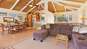 A spacious living area with a high ceiling, wooden beams, a large sofa, a dining table with chairs, and a kitchen in the background.