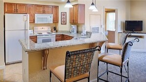 Una cocina con armarios de madera, un refrigerador blanco, un microondas y una estufa. Hay dos taburetes de bar con asientos acolchados.