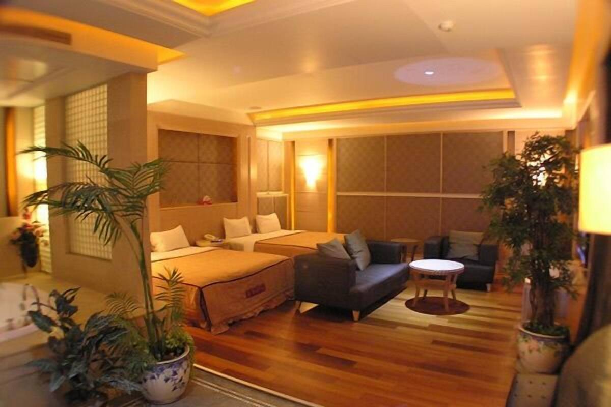 A hotel room with two beds, a sofa, a small table, and several potted plants.
