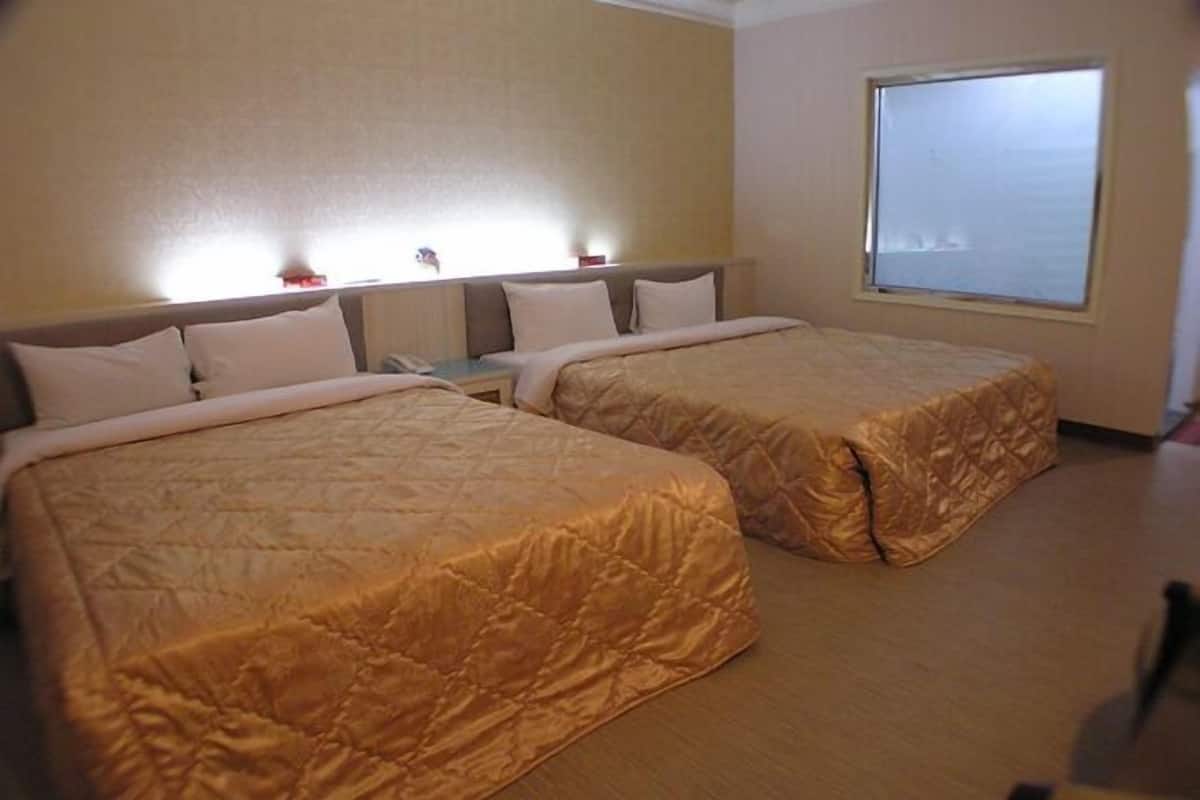 A hotel room with two beds, a nightstand, a window, and a wall-mounted light.