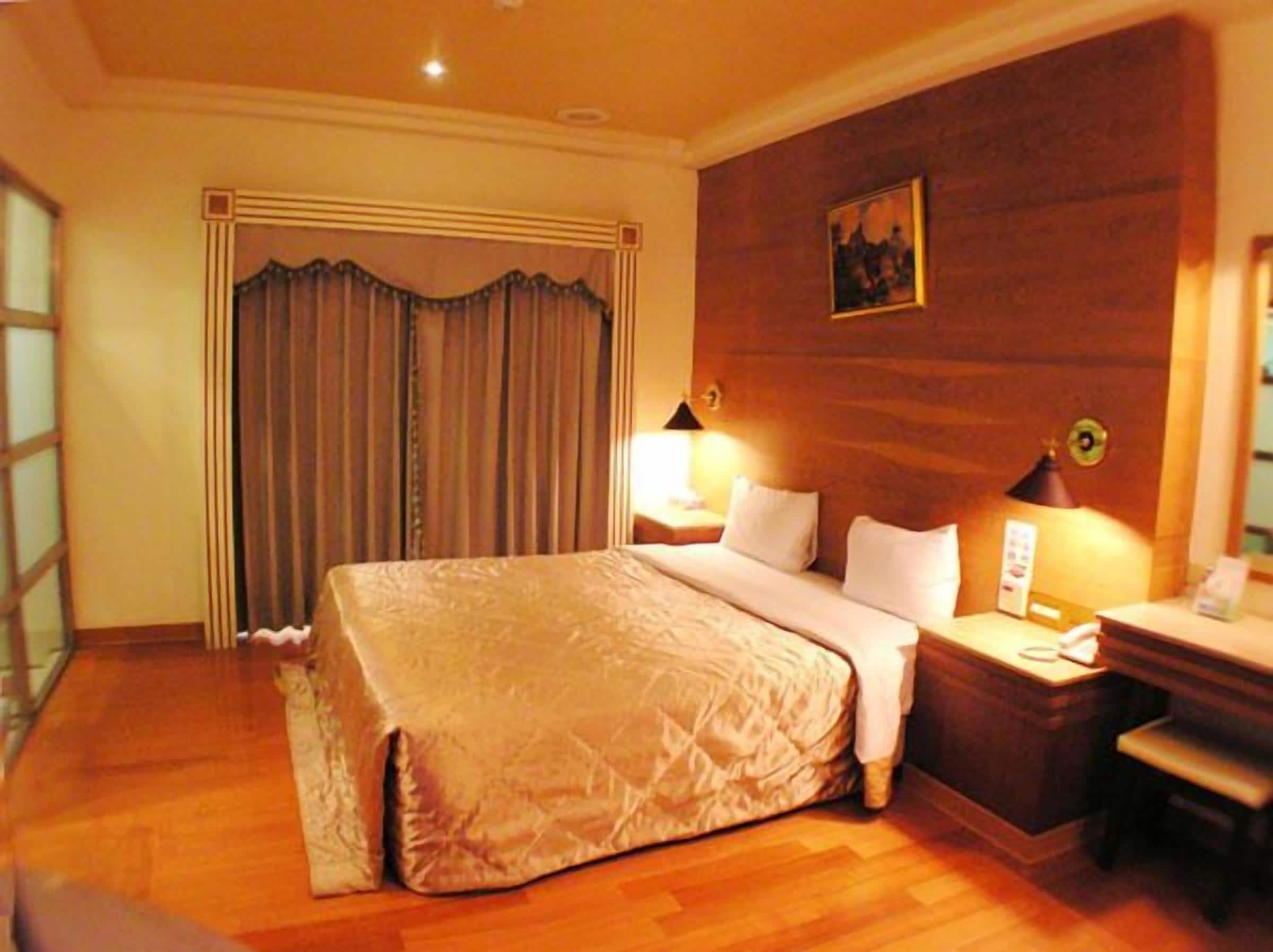 Habitación de hotel con una cama grande, cabecera de madera, dos lámparas de noche, un escritorio con una silla y un cuadro en la pared.