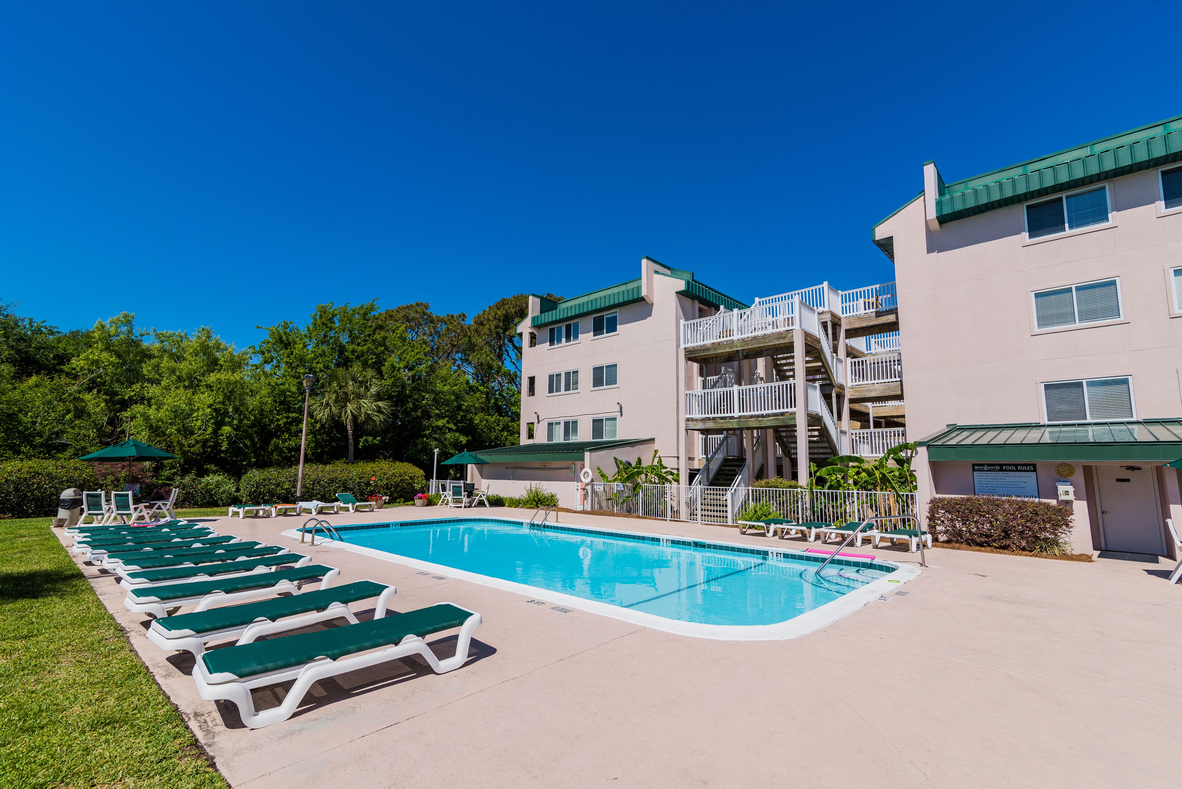 A swimming pool with lounge chairs, a staircase to a balcony, and a building with a green roof.
