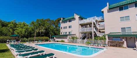 A swimming pool with lounge chairs, a staircase to a balcony, and a building with a green roof.
