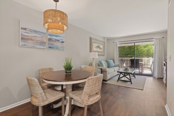 A dining area with wicker chairs, a round table, and a hanging light fixture. There is a sofa and a view of a balcony with curtains.