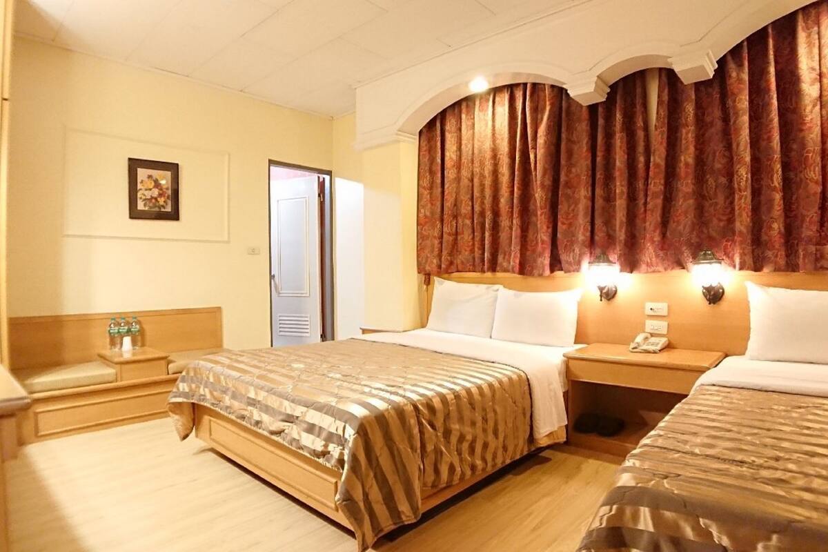 A hotel room with two beds, a bedside table with a lamp, a nightstand with a phone, and a door leading to a bathroom.