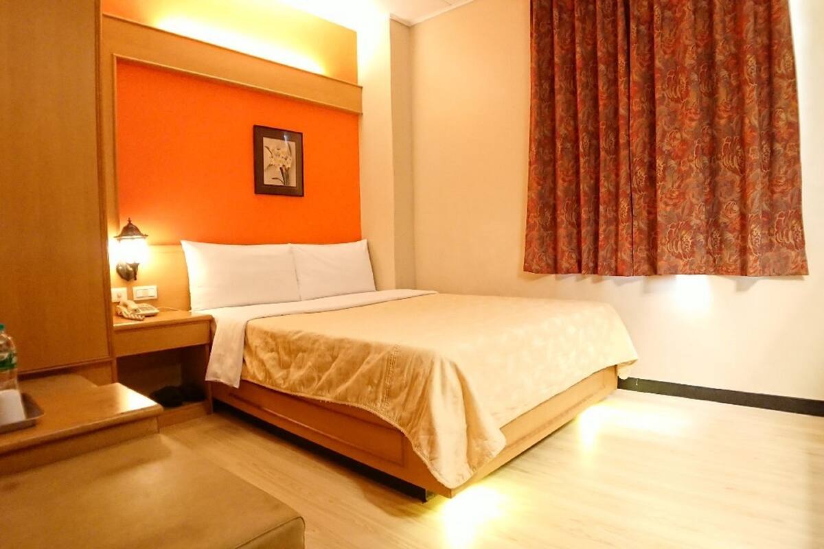 A hotel room with a bed, a nightstand with a lamp, a picture on the wall, and a window with curtains.