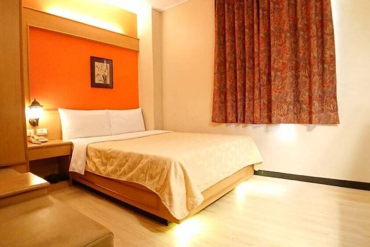 A hotel room with a bed, a nightstand, a lamp, a picture on the wall, and a window with curtains.