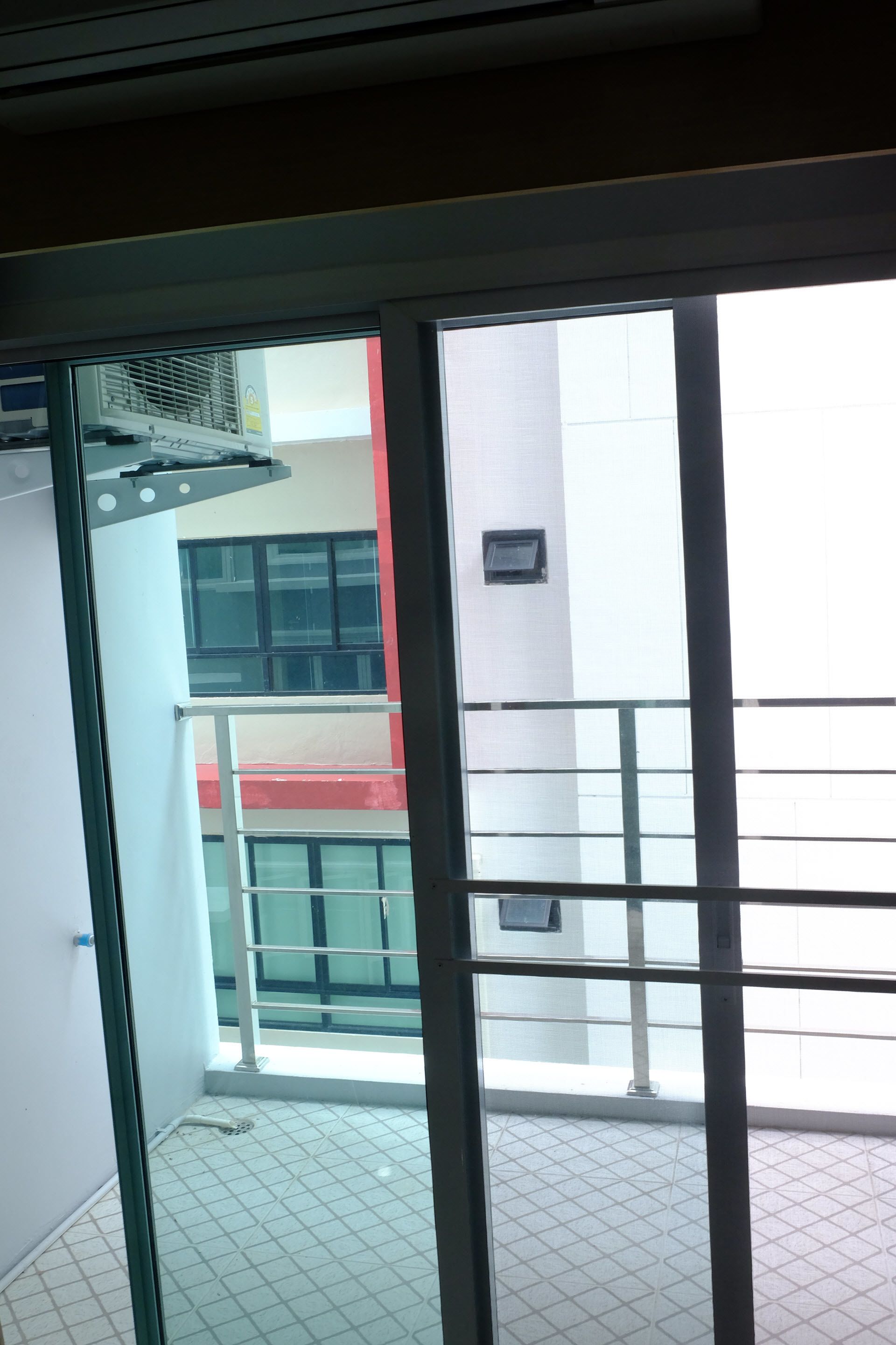 A view through a glass door into a balcony area with a tiled floor, a metal railing, and a building with a red and white facade in the background.