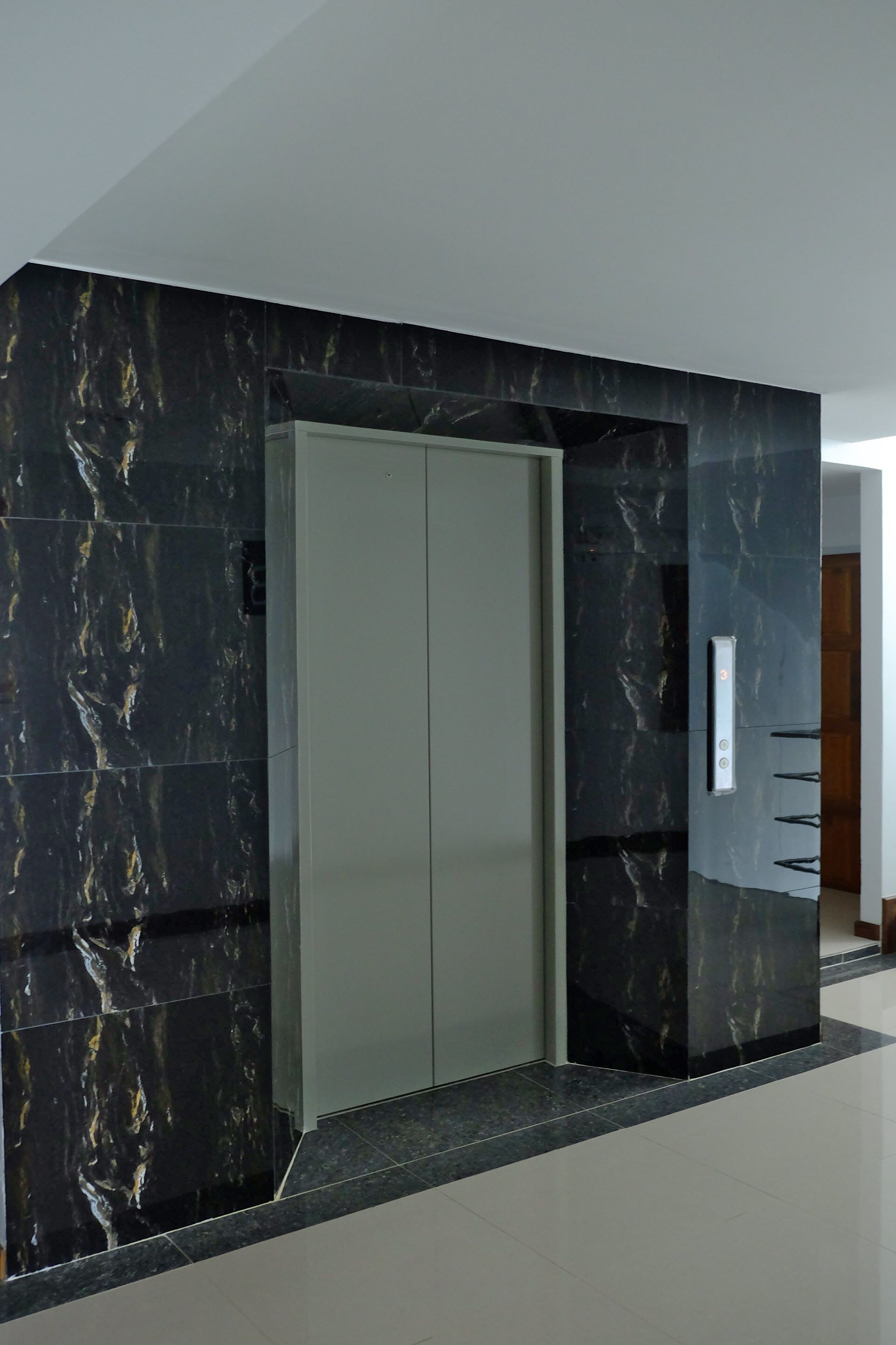 A modern elevator with a marble finish, a metallic door, and a control panel on the wall.