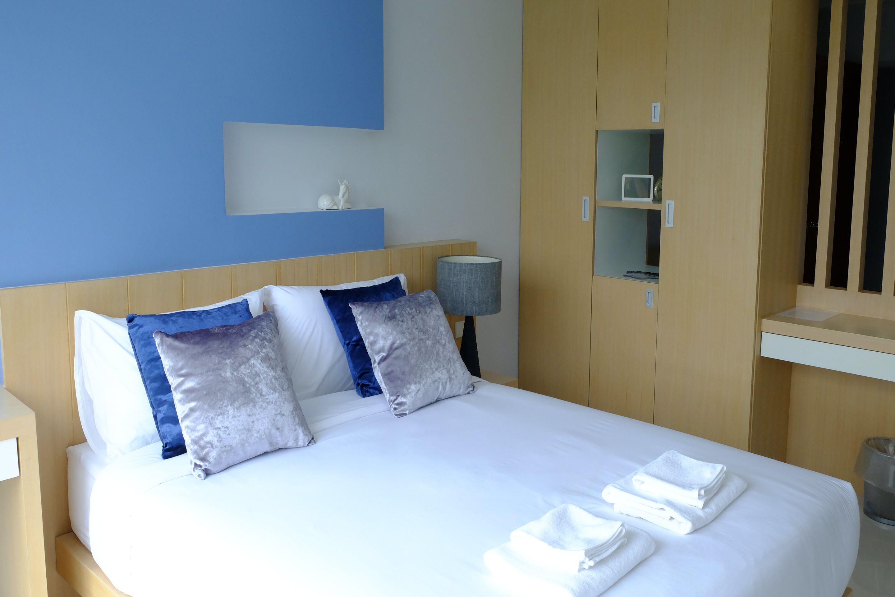 A neatly made bed with white linens and multiple pillows, a bedside lamp, and a small shelf with decorative items.