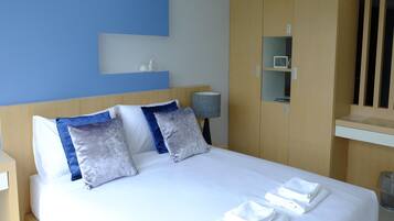 A neatly made bed with white linens and multiple pillows, a bedside lamp, and a small shelf with decorative items.
