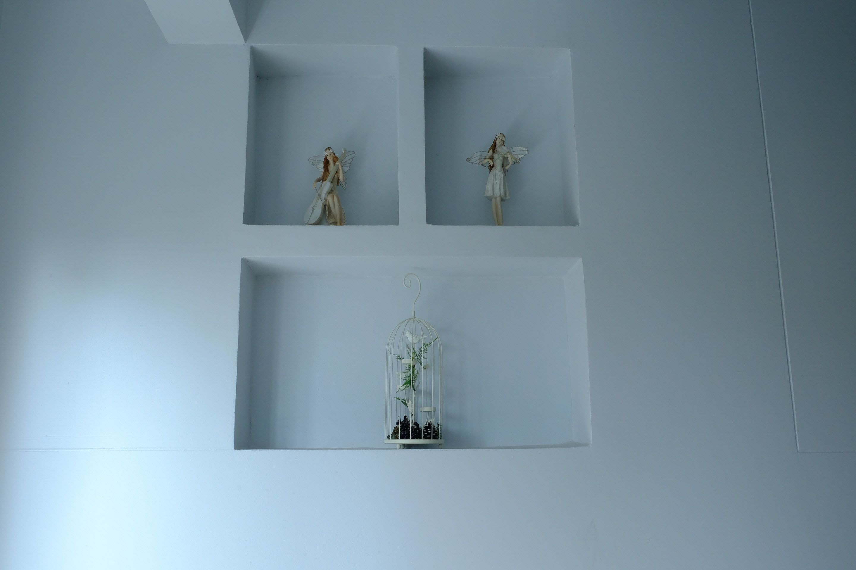 A minimalist wall with three framed items: two angel figurines and a birdcage with plants.
