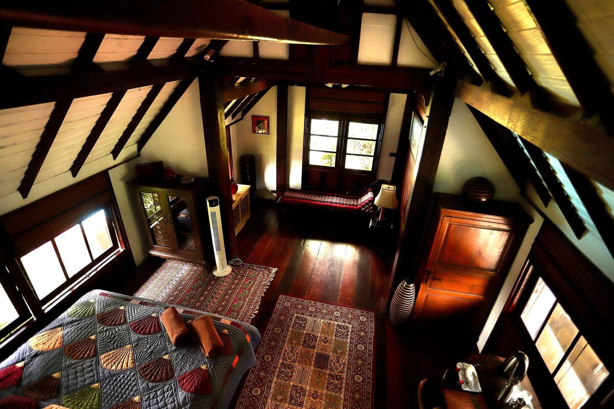 A room with a sloped ceiling, wooden beams, a bed with a quilt, a standing heater, and a window with blinds.