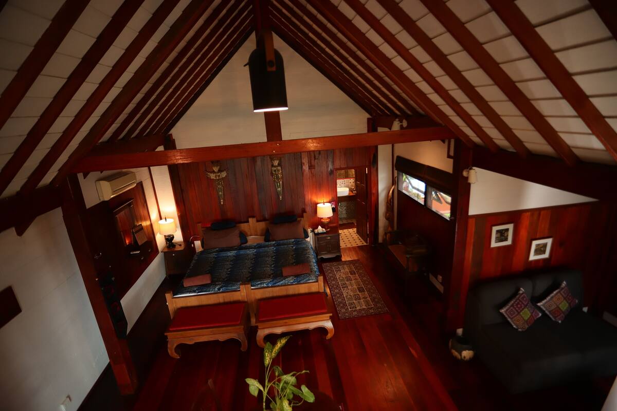 A cozy attic bedroom with a wooden ceiling, a bed with a blue bedspread, a small table with a lamp, and a sofa with cushions.